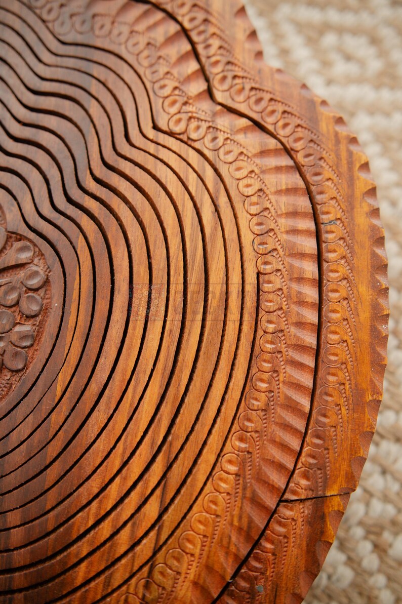 May include: Close-up of a wooden fruit basket with a carved design. The basket is made of light brown wood and features a series of curved, grooved lines. The edges are decorated with a repeating pattern of small circles and lines.