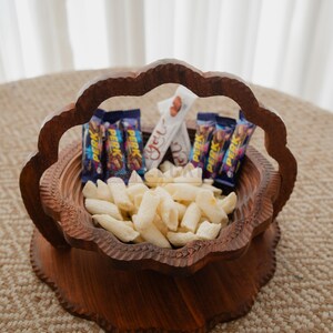 May include: A decorative wooden basket filled with snacks. The basket has a carved floral design and a handle. Inside are white snacks, chocolate bars, and a white candy bar.