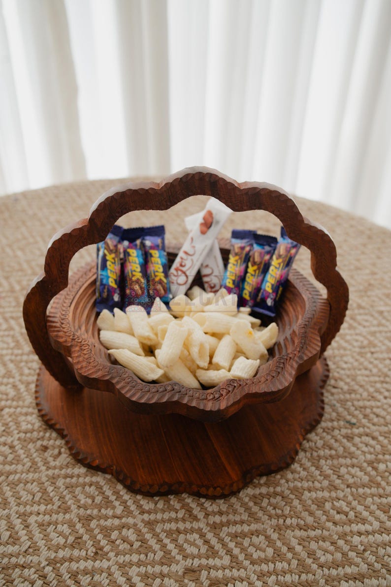 May include: A decorative wooden basket filled with snacks. The basket has a carved floral design and a handle. Inside are white snacks, chocolate bars, and a white candy bar.