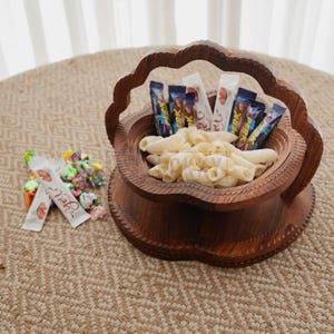 May include: A decorative wooden bowl filled with pasta and candy bars. The bowl has a carved floral design and a handle. A small pile of wrapped candies sits next to the bowl on a woven surface. The background includes a sheer curtain.