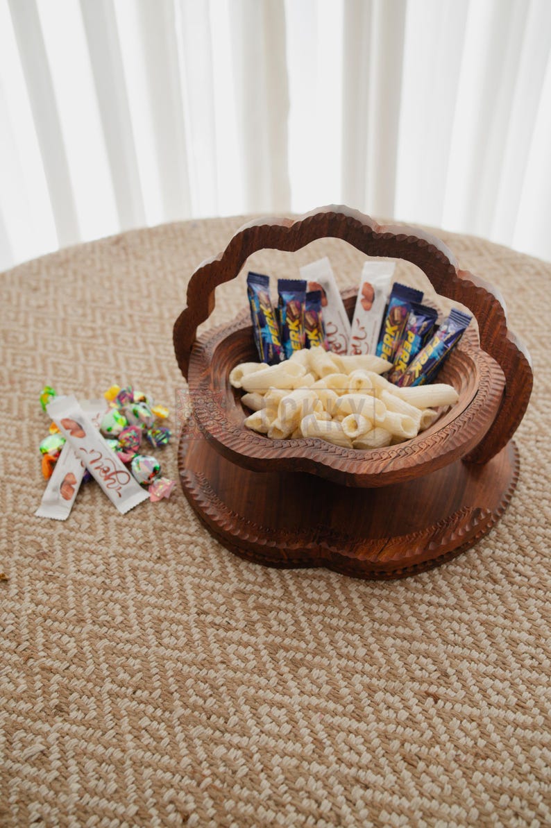 May include: A decorative wooden bowl filled with pasta and candy bars. The bowl has a carved floral design and a handle. A small pile of wrapped candies sits next to the bowl on a woven surface. The background includes a sheer curtain.
