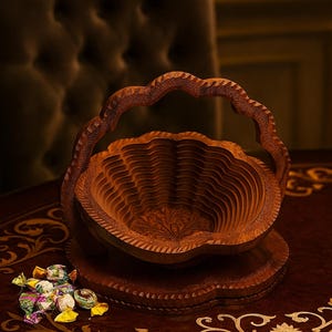 May include: A decorative wooden basket with a handle, carved from a warm brown wood. The basket has a layered design and sits on a matching base. A small pile of wrapped candies rests nearby. The basket is on a table with ornate gold detailing.