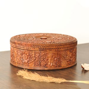 May include: A circular wooden box featuring detailed floral carvings. The box is brown and has a lid with a central knob. The sides and top are decorated with elaborate, carved floral patterns. It rests on a wooden surface.