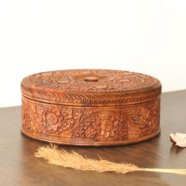 May include: A circular wooden box featuring detailed floral carvings. The box is brown and has a lid with a central knob. The sides and top are decorated with elaborate, carved floral patterns. It rests on a wooden surface.