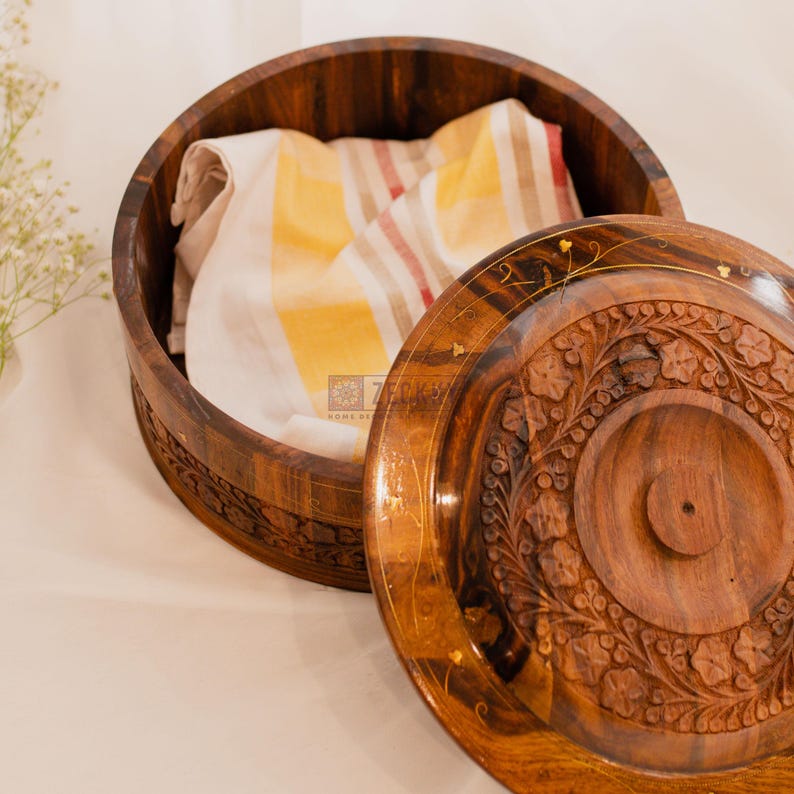 May include: A round, wooden storage box with a lid. The box is made of dark brown wood and features carved floral designs. Inside the box is a folded white and yellow striped cloth. The lid is partially open, revealing the contents.