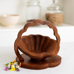May include: A dark brown, wooden fruit basket with a carved handle and base. The basket has a scalloped edge and a folding design. A small pile of colorful candies sits in front of the basket.