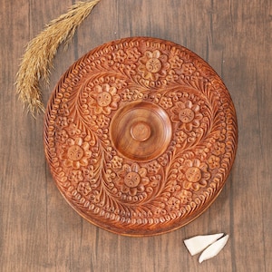 May include: A round, intricately carved wooden serving tray with a floral design. The tray is brown and features a central recessed area and a detailed border. A sprig of dried grass and two white pieces are next to the tray.