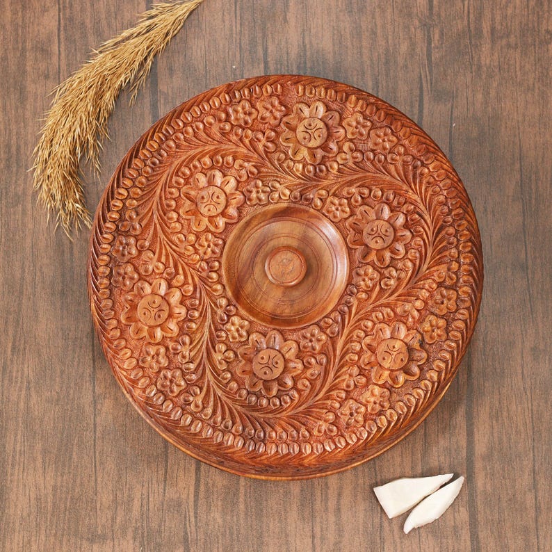 May include: A round, intricately carved wooden serving tray with a floral design. The tray is brown and features a central recessed area and a detailed border. A sprig of dried grass and two white pieces are next to the tray.