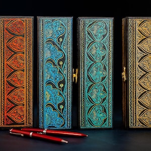 Pode incluir: Quatro estojos retangulares para canetas com designs coloridos e ornamentados. Os estojos são pretos com fechos dourados e apresentam padrões em laranja, verde-azulado e dourado. Duas canetas vermelhas repousam em frente aos estojos, sugerindo um conjunto de escrita ou presente.