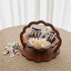 May include: A decorative wooden serving bowl filled with snacks and candy. The bowl has a unique scalloped edge design and is filled with white snacks and wrapped candies. Several snack bars are also included.