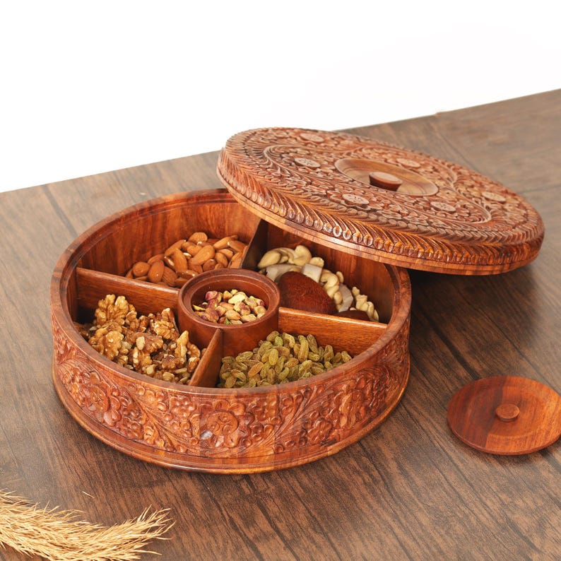 May include: A circular wooden serving platter with intricate floral carvings. It's divided into five compartments, containing almonds, walnuts, pistachios, raisins, and other nuts. The lid is ajar, displaying the contents. The platter rests on a wooden surface.