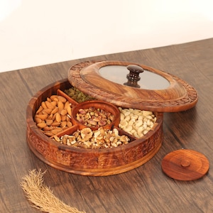 May include: A circular wooden serving platter with a glass-topped lid and a wooden handle. The tray is divided into compartments containing almonds, pistachios, walnuts, cashews, and raisins. The tray is on a wooden surface.
