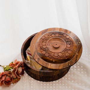 May include: A wooden round box with a lid, featuring intricate floral carvings. The box is open, revealing a glimpse of the interior. The lid is partially lifted, showcasing the detailed craftsmanship and the natural wood grain. The box is a warm brown color.