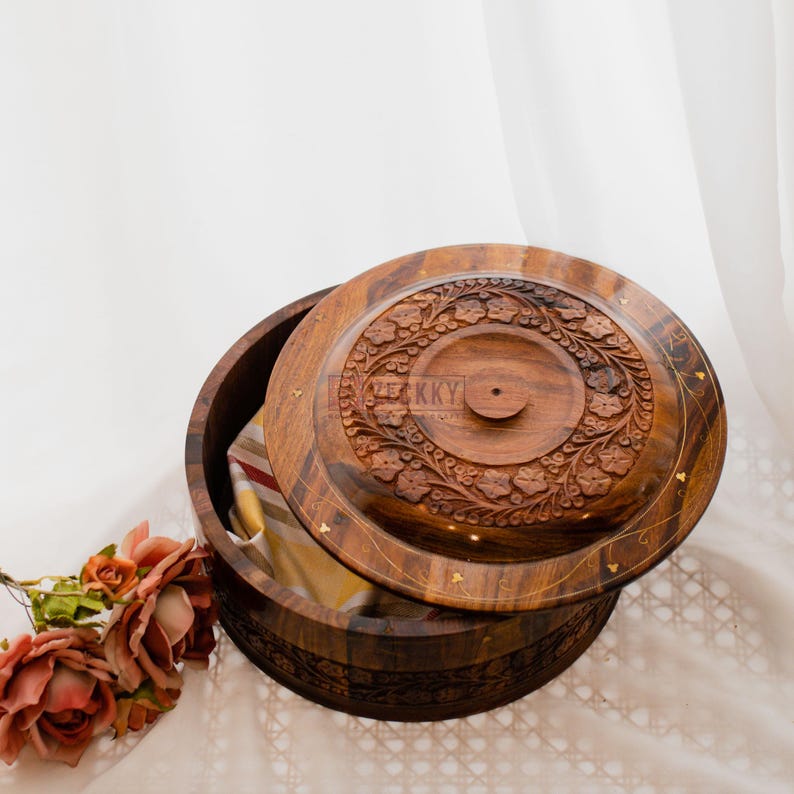 May include: A wooden round box with a lid, featuring intricate floral carvings. The box is open, revealing a glimpse of the interior. The lid is partially lifted, showcasing the detailed craftsmanship and the natural wood grain. The box is a warm brown color.