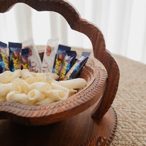 May include: A handcrafted wooden serving bowl with a decorative handle, filled with snacks and candy bars. The bowl has intricate carvings and a rich, dark brown finish. The snacks include white pasta-shaped chips and various wrapped chocolate bars.