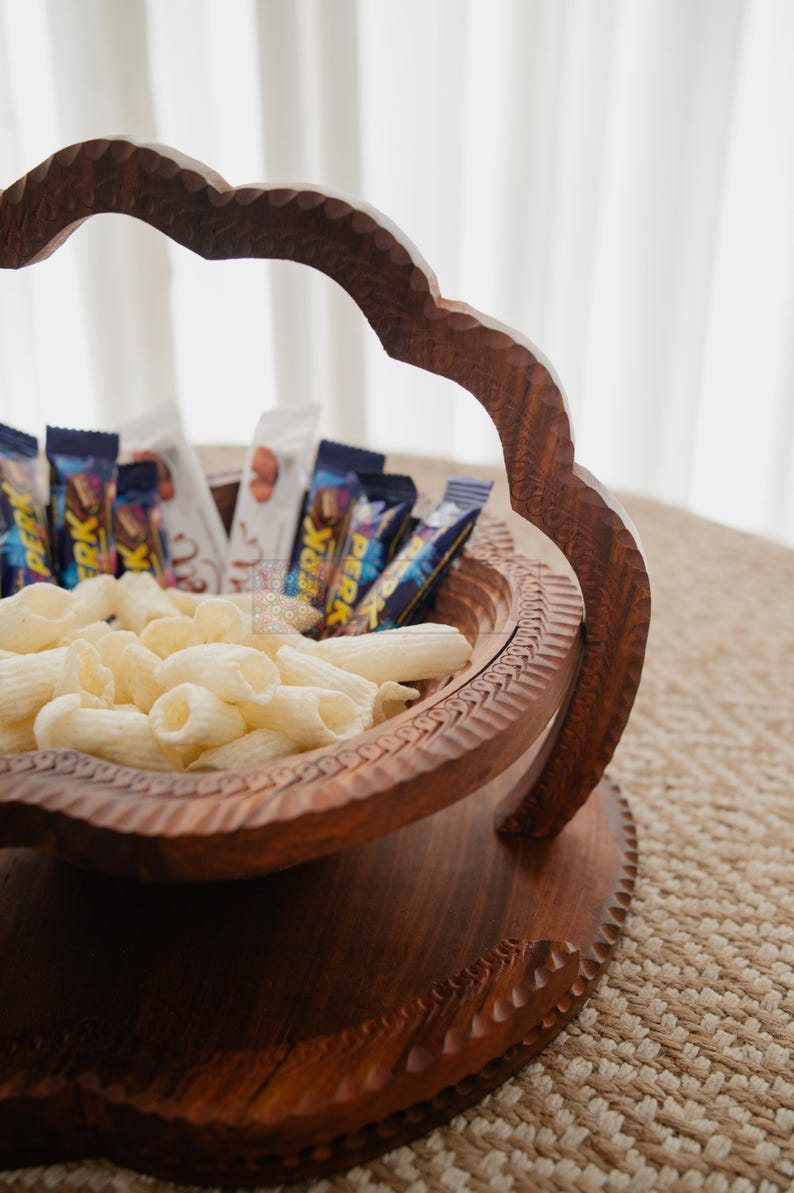 May include: A handcrafted wooden serving bowl with a decorative handle, filled with snacks and candy bars. The bowl has intricate carvings and a rich, dark brown finish. The snacks include white pasta-shaped chips and various wrapped chocolate bars.