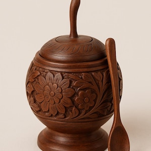 Puede incluir: Un azucarero de madera marrón oscuro con tapa y cuchara a juego. El cuenco es redondo con diseños florales tallados. La tapa tiene un asa curvada. La cuchara es larga y delgada. El juego es de madera.