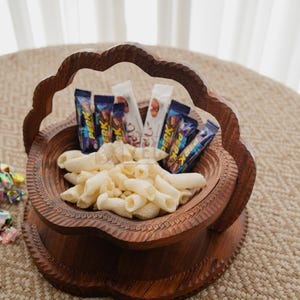 May include: A decorative wooden basket filled with penne pasta and assorted candy bars. The basket has a carved design and a handle. The contents are arranged on a woven surface.