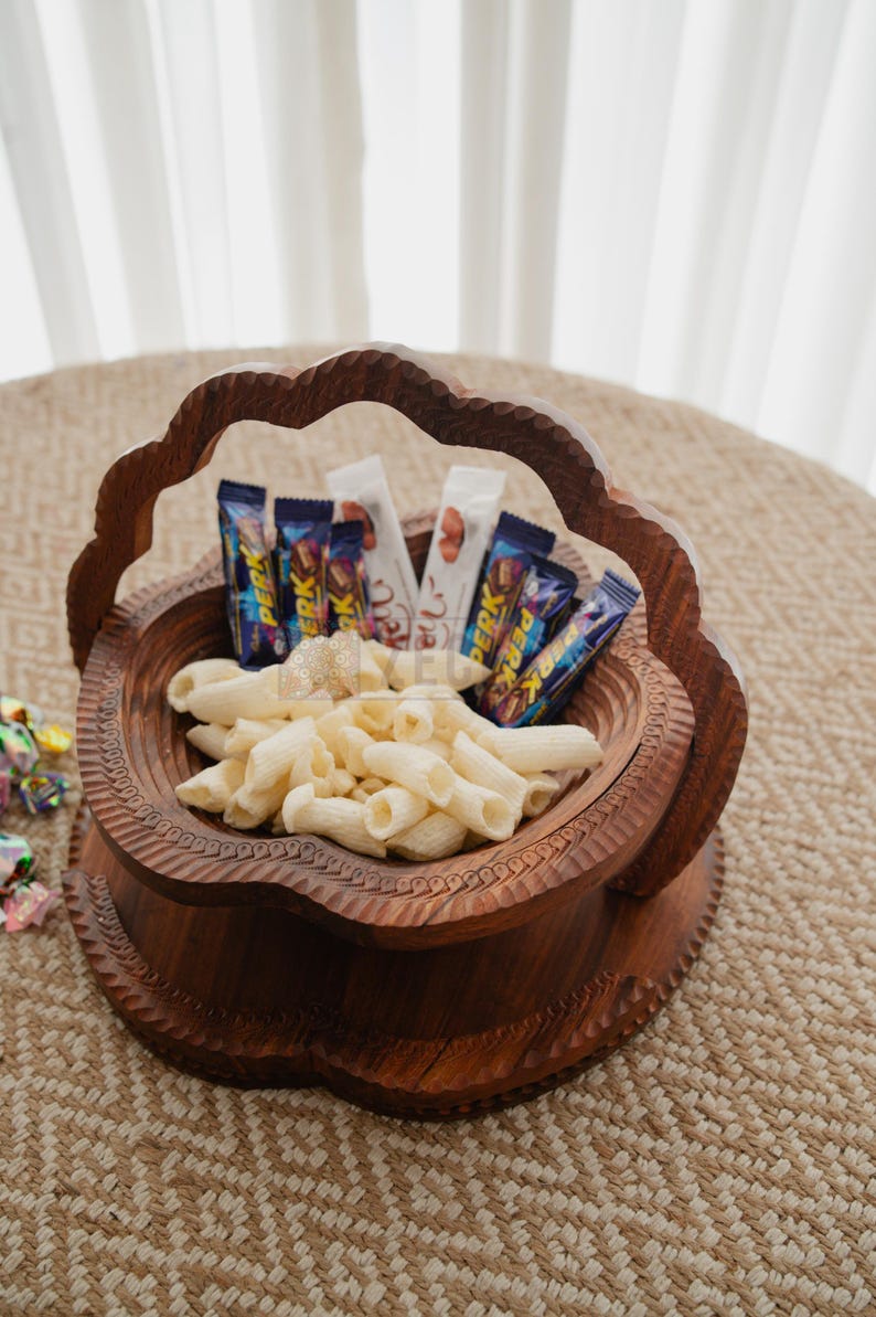 May include: A decorative wooden basket filled with penne pasta and assorted candy bars. The basket has a carved design and a handle. The contents are arranged on a woven surface.