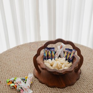 May include: A wooden serving bowl filled with snacks and candies. The bowl has a unique, scalloped edge design and is filled with white, puffed snacks and small candy bars. A small pile of wrapped candies sits nearby.