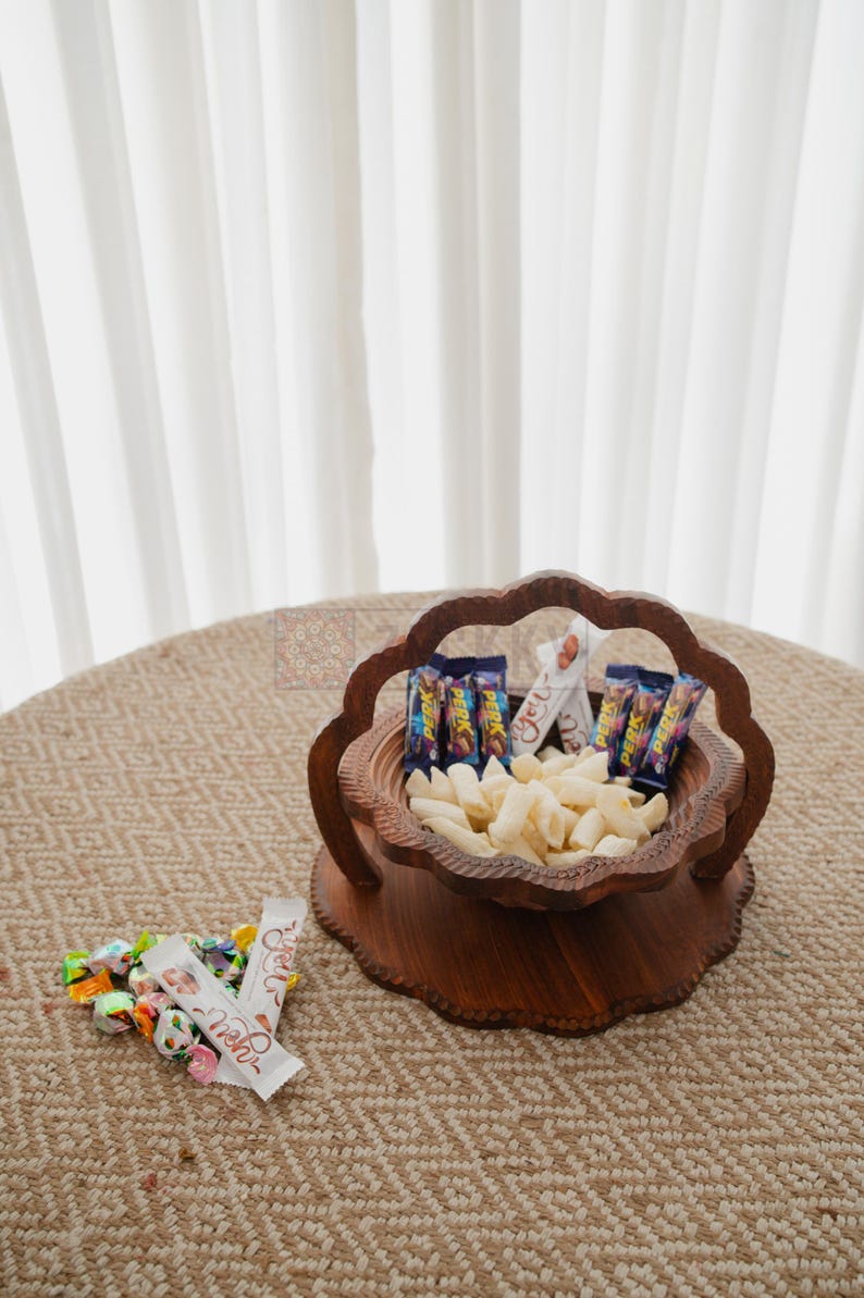 May include: A wooden serving bowl filled with snacks and candies. The bowl has a unique, scalloped edge design and is filled with white, puffed snacks and small candy bars. A small pile of wrapped candies sits nearby.