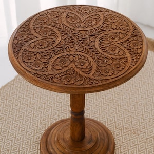 May include: A round, wooden side table with intricate floral carvings on the tabletop. The table has a single pedestal base and a warm, brown finish. The table is approximately 20 inches in diameter and 24 inches tall.