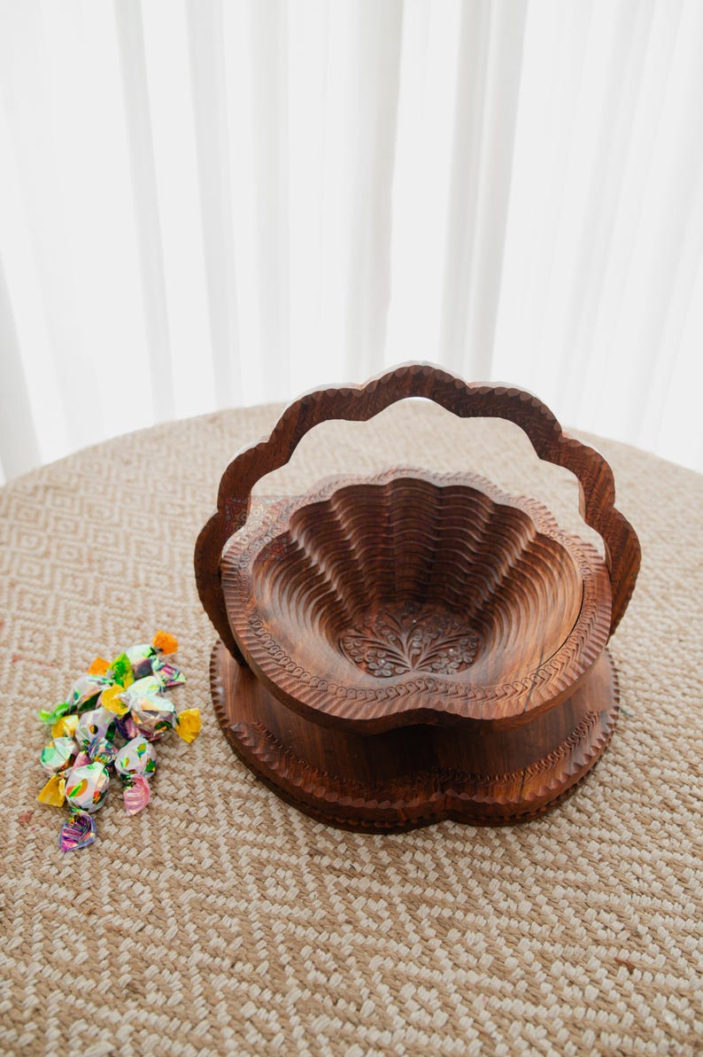 May include: A carved wooden fruit basket with a scalloped edge and handle. The basket is brown and sits on a round base. A small pile of wrapped candies in various colors is next to the basket.