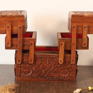 May include: A dark brown wooden jewellery box with a multi-tiered design. The box features detailed floral carvings and a red fabric lining. It has multiple compartments and hinged lids. The box is designed to store jewellery.