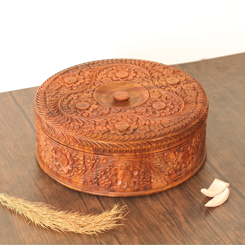 May include: A circular wooden box, intricately carved with floral patterns. The box is a warm brown hue and has a lid with a central knob. The detailed carvings adorn the top and sides, displaying a handcrafted design. The box is placed on a wooden surface.
