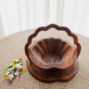May include: A carved wooden fruit basket with a scalloped edge and handle. The basket is brown and sits on a round base. A small pile of wrapped candies in various colors is next to the basket.