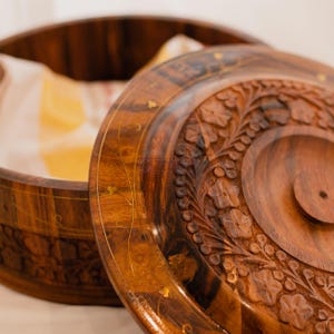 May include: A partially open, intricately carved wooden box with a lid. The box is a warm brown color with floral and leaf patterns carved into the lid. The rim of the box and lid are decorated with small gold accents.