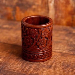 Puede incluir: Un recipiente cilíndrico de madera marrón oscuro con patrones florales tallados. El recipiente presenta tallas en relieve detalladas de flores y hojas alrededor de su exterior, con un borde decorativo en la base. El interior es liso y vacío.
