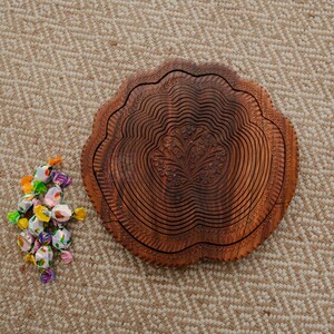May include: A decorative wooden fruit bowl with a carved floral design. The bowl is brown and has a unique, layered design. A small pile of wrapped candies in various colors sits next to the bowl on a patterned beige surface.