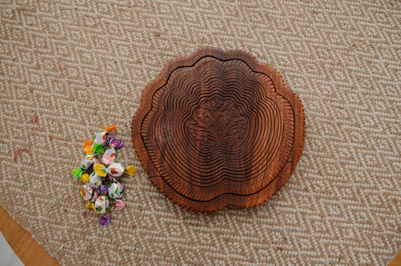 May include: A decorative wooden fruit bowl with a carved floral design. The bowl is brown and has a unique, layered design. A small pile of wrapped candies in various colors sits next to the bowl on a patterned beige surface.