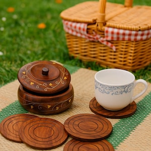 May include: A picnic scene featuring a woven basket with a red and white checkered lining, a wooden container with a lid, a white teacup, and several wooden coasters. The items are arranged on a woven mat.