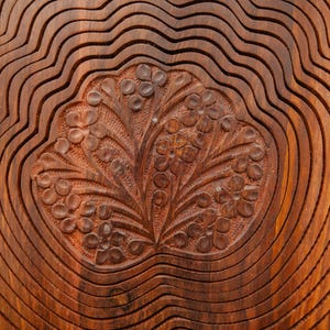 May include: Close-up of a wooden decorative bowl with a carved floral design. The bowl has a warm brown color and a wavy, layered pattern. The floral carving is in the center, adding a touch of elegance to the wooden bowl.