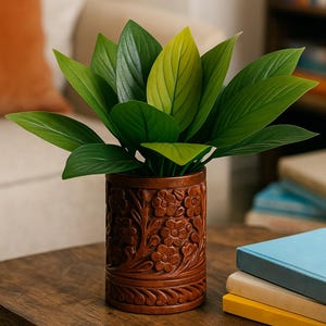 May include: A decorative wooden planter with carved floral designs holds a vibrant green plant. The leaves are a mix of light and dark green. The planter sits on a wooden table, with books in the background.