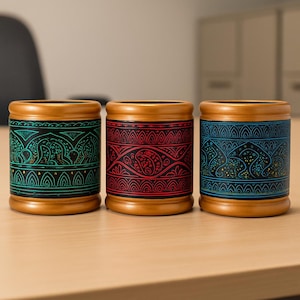 Puede incluir: Tres recipientes cilíndricos con diseños intrincados y coloridos. Los recipientes tienen una base y un borde de madera, con el cuerpo principal con patrones en verde azulado, rojo y azul. Los diseños incluyen elementos florales y geométricos.