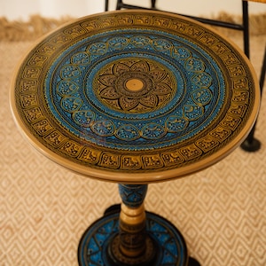 May include: A round side table with a decorative, hand-painted design. The tabletop features a central floral motif in blue and gold, surrounded by intricate patterns. The table has a pedestal base and measures approximately 20 inches in diameter.