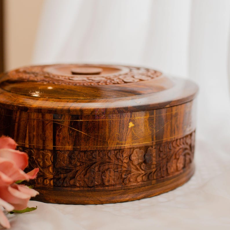 May include: A round, wooden box with a lid. The box is a rich brown color and features intricate floral carvings around the sides and on the lid. The lid has a small, raised handle. The box is sitting on a white surface.