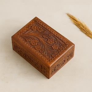 Puede incluir: Una caja rectangular de madera con intrincadas tallas florales. La caja es de color marrón cálido y presenta patrones en relieve detallados en la tapa y los lados. El diseño incluye flores y vides arremolinadas, creando un artículo de almacenamiento decorativo.
