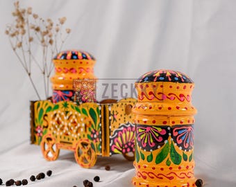 May include: A decorative, hand-painted wooden cart and two matching spice shakers. The cart and shakers are painted in bright yellow with intricate floral and geometric designs in green, pink, and black. The shakers have a rounded top.