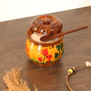 May include: A wooden salt cellar with a lid and a small spoon. The yellow container is decorated with red flowers and green trees. The lid is slightly ajar, revealing the salt inside.