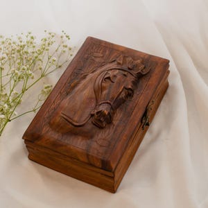 Könnte beinhalten: Rechteckige Holzbox mit detaillierter Reliefschnitzerei eines Pferdekopfes. Die Box hat eine warme braune Farbe und einen Klappdeckel mit Metallverschluss. Die Pferdeschnitzerei ist detailliert und zeigt Zaumzeug und Gesichtszüge.