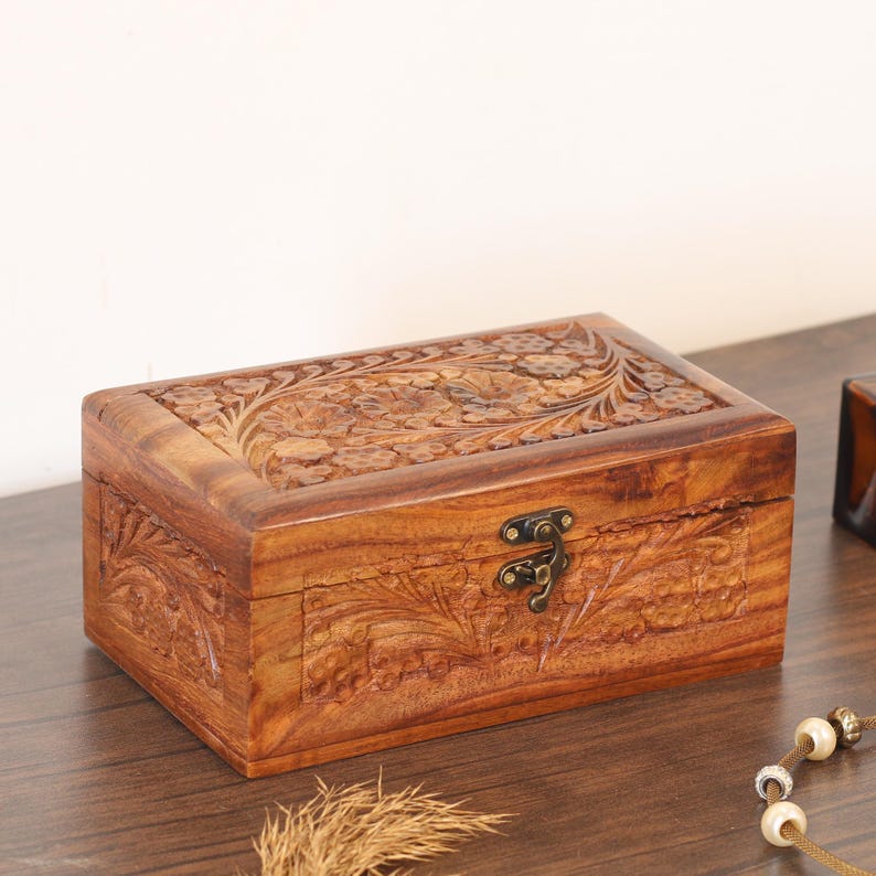 Op de afbeelding: Een rechthoekige houten kist met ingewikkelde bloemensnijwerk op het deksel en de zijkanten. De kist heeft een warme bruine kleur en een metalen sluiting. Ideaal voor het opbergen van sieraden of kleine aandenkens.