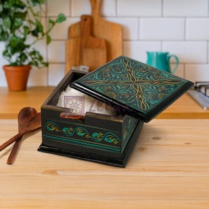 Puede incluir: Caja de madera negra con tapa abierta, con diseños florales turquesa y dorados. La caja está decorada con diseños ondulados turquesa y dorados. Dentro de la caja hay una pequeña tarjeta estampada. La caja está sobre una superficie de madera.