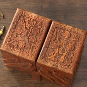 Puede incluir: Dos cajas de madera con intrincadas tallas florales. Las cajas son de color marrón cálido y parecen estar unidas por bisagras. El diseño floral cubre la parte superior y los lados de las cajas, creando un elemento de almacenamiento decorativo.