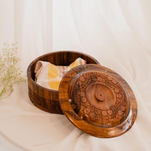 May include: A round, open wooden box with a carved floral design on the lid. The lid is partially open, revealing a folded cloth inside. The box and lid are a warm brown color, with small brass accents.
