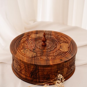 Könnte beinhalten: Eine runde Holzbox mit Deckel, verziert mit aufwendigen Blumenschnitzereien und einem kleinen Knopf oben. Die Box hat eine warme braune Farbe und scheint aus Holz zu sein. Sie liegt auf einer weißen Oberfläche.