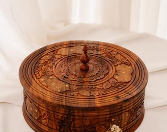 May include: A round, wooden box with a lid, featuring intricate floral carvings and a small knob on top. The box is a warm brown color and appears to be made of wood. The box is sitting on a white surface.